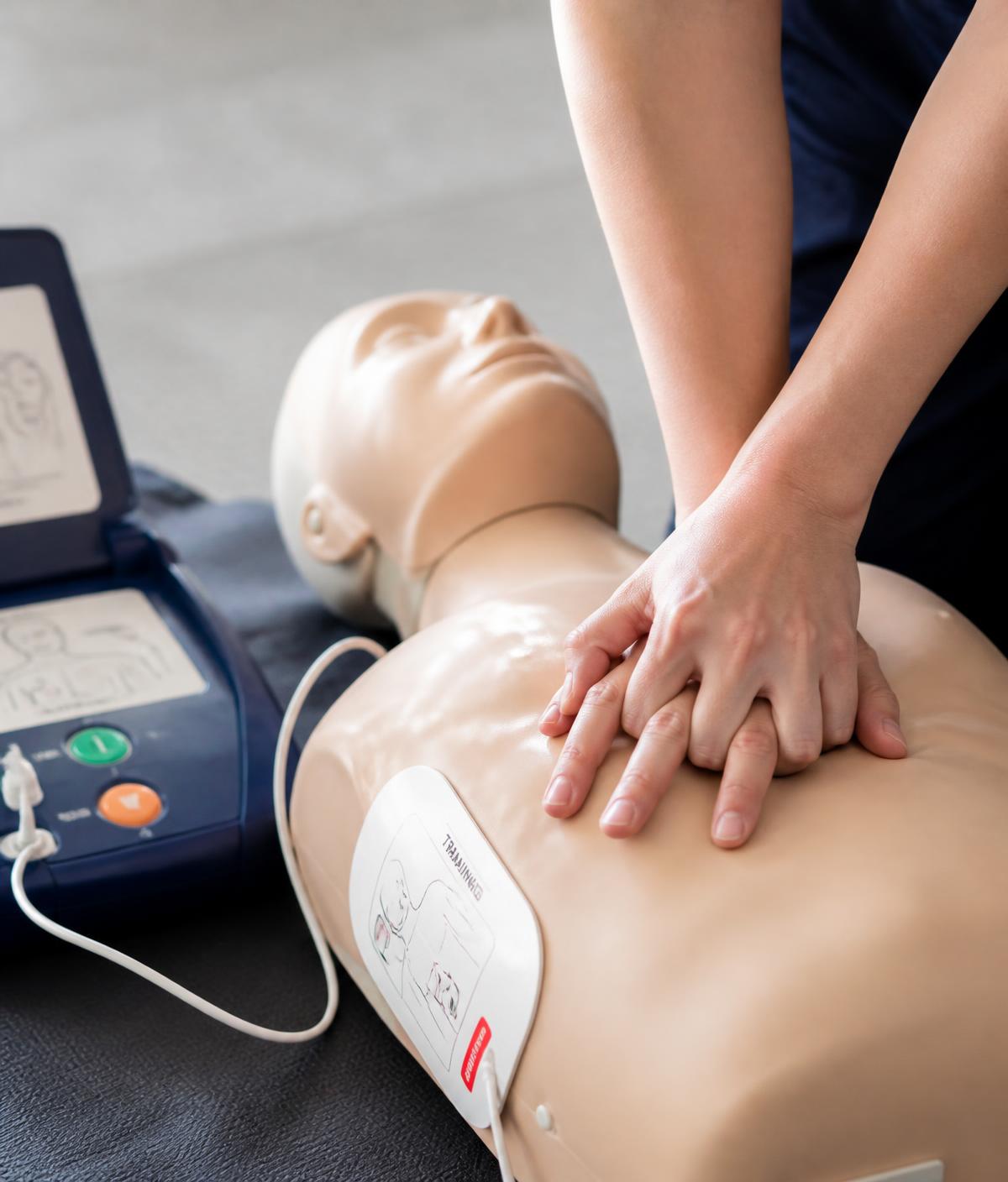 Close-up of hands performing chest compressions on a training manikin beside an AED